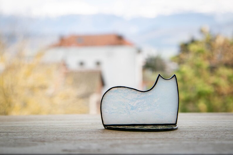 May include: A stained glass cat figurine featuring a white and pale blue marbled pattern with a smooth, curved silhouette. The piece is outlined in a dark, thin metal solder frame that defines the shape of the cat's back, ears, and base.