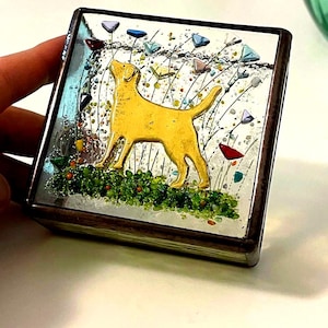 May include: A square, decorative trinket box with a yellow Labrador Retriever design. The box features a mirrored surface with colorful accents, including green grass, and multi-colored triangular shapes. The box has a dark brown frame.