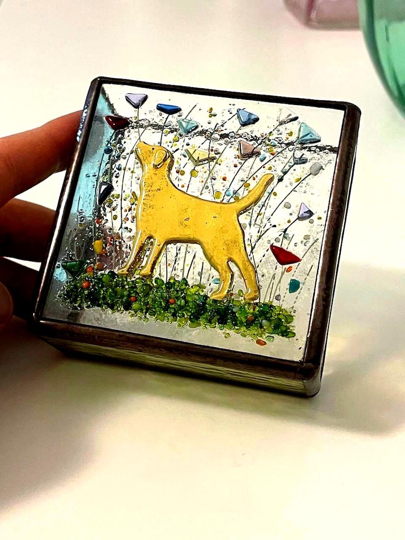 May include: A square, decorative trinket box with a yellow Labrador Retriever design. The box features a mirrored surface with colorful accents, including green grass, and multi-colored triangular shapes. The box has a dark brown frame.