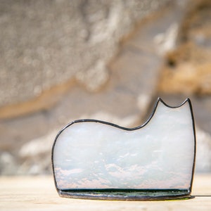 May include: A stained glass cat figurine featuring a smooth, translucent white body with subtle iridescent highlights. The cat shape is outlined in a dark, thin metal solder frame, creating a clean silhouette that rests on a flat base.