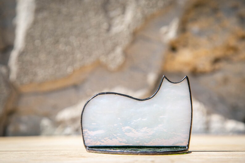 May include: A stained glass cat figurine featuring a smooth, translucent white body with subtle iridescent highlights. The cat shape is outlined in a dark, thin metal solder frame, creating a clean silhouette that rests on a flat base.