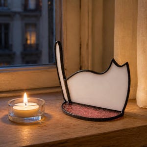 May include: A white stained glass cat silhouette with a dark metal frame and a textured pink glass base, positioned next to a small, round, clear glass tea light candle holder containing a lit white wax candle with a visible yellow flame.
