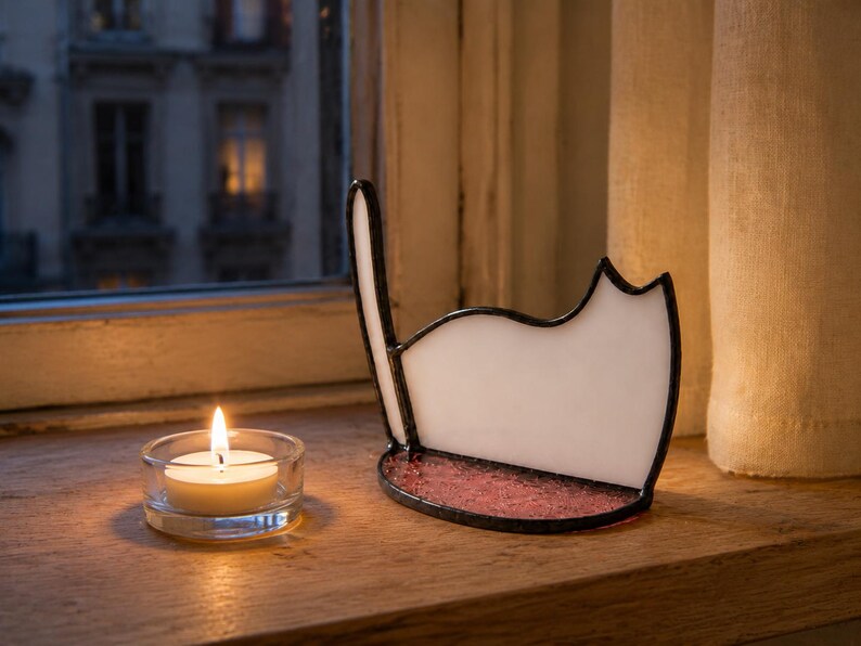 May include: A white stained glass cat silhouette with a dark metal frame and a textured pink glass base, positioned next to a small, round, clear glass tea light candle holder containing a lit white wax candle with a visible yellow flame.