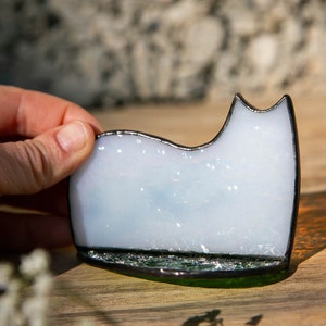 May include: A white stained glass cat silhouette with a subtle iridescent finish and a dark soldered metal edge. The cat figure is mounted on a dark green glass base, creating a smooth, reflective surface that catches the light.