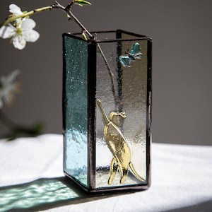 May include: A rectangular glass vase with a teal and clear color scheme. A gold-colored cat and a blue butterfly are affixed to the glass. A branch with white flowers is inside the vase.