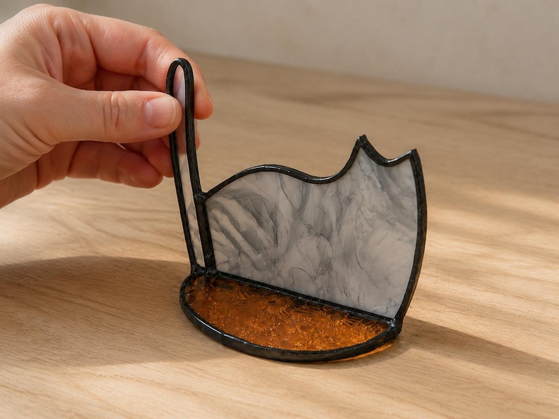 May include: A stained glass cat-shaped stand featuring a white and gray marbled body with a dark metal frame. The base is made of textured amber-colored glass, and the structure includes a vertical tail piece extending upward from the side.