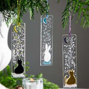May include: Three glass baubles with a white crescent moon and a different animal on each. The baubles are hanging from a green tree branch. The animals are a black cat, a white rabbit, and a gold cat.