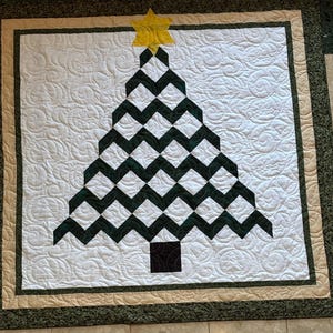May include: A square quilt featuring a central Christmas tree design composed of dark green and white chevron-patterned fabric blocks. A yellow star sits atop the tree, and a dark square forms the base. The white background has a swirling quilted texture.