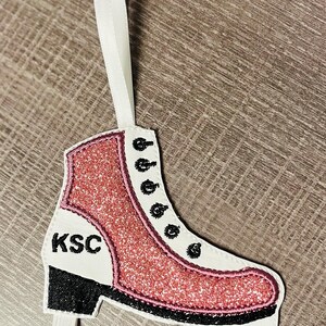 K&ouml;nnte beinhalten: Rosa glitzernder Schlittschuh-Anh&auml;nger mit schwarzem Besatz und den Buchstaben "KSC" an der Seite. Der Anh&auml;nger hat ein wei&szlig;es Band zum Aufh&auml;ngen.