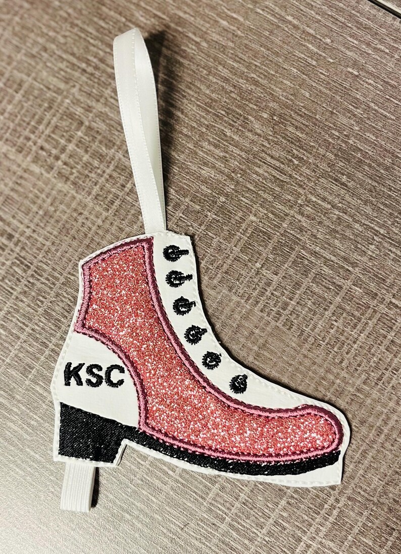 K&ouml;nnte beinhalten: Rosa glitzernder Schlittschuh-Anh&auml;nger mit schwarzem Besatz und den Buchstaben "KSC" an der Seite. Der Anh&auml;nger hat ein wei&szlig;es Band zum Aufh&auml;ngen.