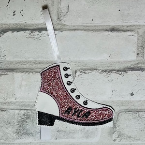 K&ouml;nnte beinhalten: Rosa glitzernder Schlittschuh-Anh&auml;nger mit dem Namen "Ayla" bestickt auf der Seite. Der Anh&auml;nger ist wei&szlig; mit schwarzem Besatz und hat ein wei&szlig;es Band zum Aufh&auml;ngen.