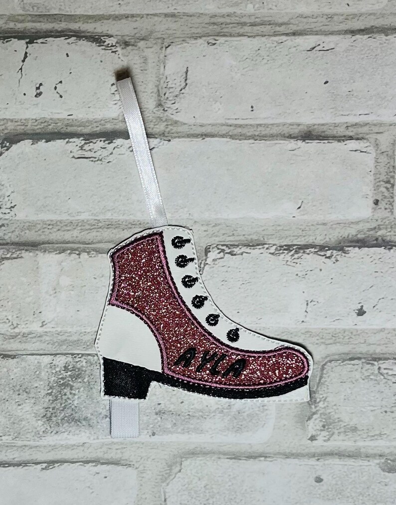 K&ouml;nnte beinhalten: Rosa glitzernder Schlittschuh-Anh&auml;nger mit dem Namen "Ayla" bestickt auf der Seite. Der Anh&auml;nger ist wei&szlig; mit schwarzem Besatz und hat ein wei&szlig;es Band zum Aufh&auml;ngen.