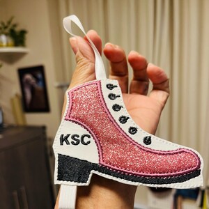 K&ouml;nnte beinhalten: Rosa glitzernder Schlittschuh-Anh&auml;nger mit schwarzen N&auml;hten und den gestickten Initialen "KSC" an der Seite.