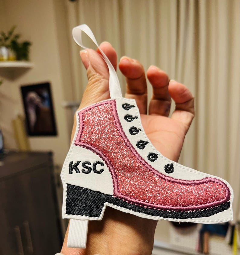 K&ouml;nnte beinhalten: Rosa glitzernder Schlittschuh-Anh&auml;nger mit schwarzen N&auml;hten und den gestickten Initialen "KSC" an der Seite.
