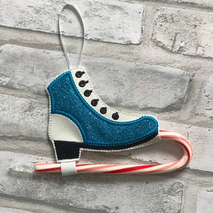 K&ouml;nnte beinhalten: Ein blauer und wei&szlig;er Schlittschuh-Anh&auml;nger mit einem Zuckerstangen-Anh&auml;nger. Der Anh&auml;nger ist mit schwarzen N&auml;hten und einem wei&szlig;en Band zum Aufh&auml;ngen verziert.