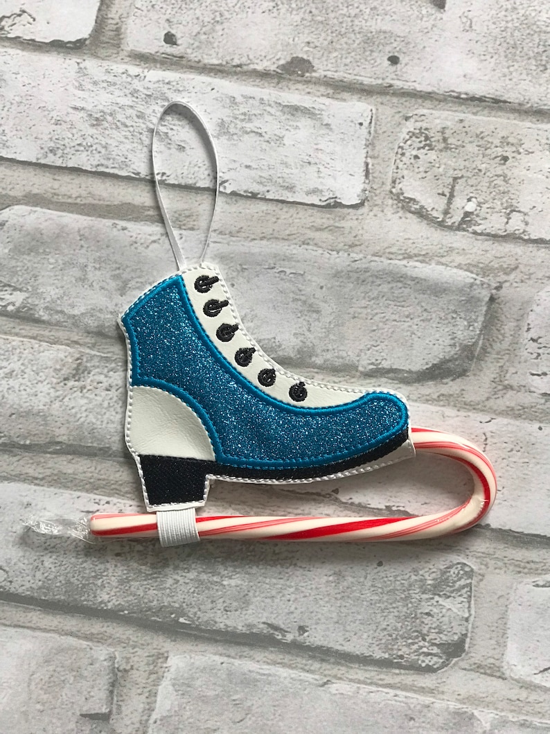 K&ouml;nnte beinhalten: Ein blauer und wei&szlig;er Schlittschuh-Anh&auml;nger mit einem Zuckerstangen-Anh&auml;nger. Der Anh&auml;nger ist mit schwarzen N&auml;hten und einem wei&szlig;en Band zum Aufh&auml;ngen verziert.