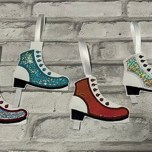 K&ouml;nnte beinhalten: Drei wei&szlig;e Filz-Schlittschuh-Anh&auml;nger mit Glitzer-Akzenten. Die Schlittschuhe sind mit rotem, blauem und orangefarbenem Glitzer verziert. Jeder Schlittschuh hat ein wei&szlig;es Band zum Aufh&auml;ngen.