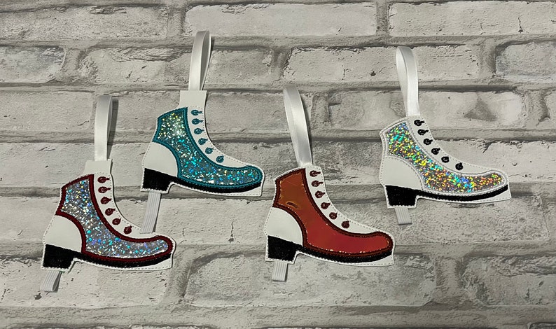 K&ouml;nnte beinhalten: Drei wei&szlig;e Filz-Schlittschuh-Anh&auml;nger mit Glitzer-Akzenten. Die Schlittschuhe sind mit rotem, blauem und orangefarbenem Glitzer verziert. Jeder Schlittschuh hat ein wei&szlig;es Band zum Aufh&auml;ngen.