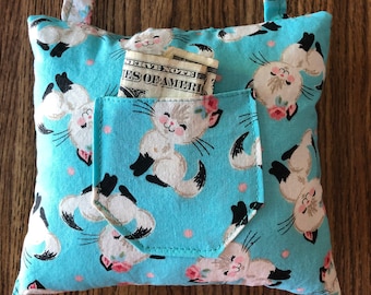 Op de afbeelding: Een klein blauw kussen met een wit kattenpatroon en een zakje aan de voorkant. Het kussen heeft een lusvormig handvat en een dollarbiljet zit in het zakje.