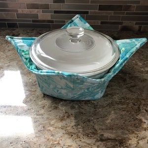 Puede incluir: Un porta ollas de tela con estampado floral azul y blanco con una cacerola de vidrio con tapa en su interior.