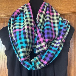 May include: A black and white plaid scarf with pink, teal, and light green accents. The scarf is folded and draped over a black shirt.
