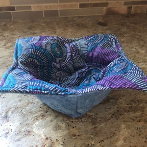 May include: A blue and purple fabric bowl cozy with a geometric pattern. The cozy is made of quilted fabric and has a square shape.