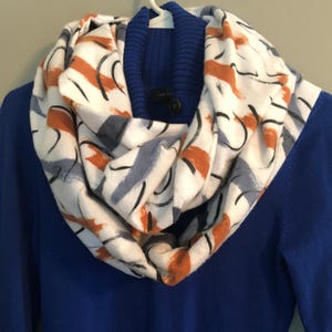 May include: A white infinity scarf with a brown, gray, and black abstract pattern. The scarf is worn over a blue sweater.