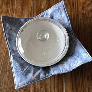 Puede incluir: Una almohadilla caliente acolchada azul con un patrón de mariposa y una tapa de vidrio transparente en la parte superior.