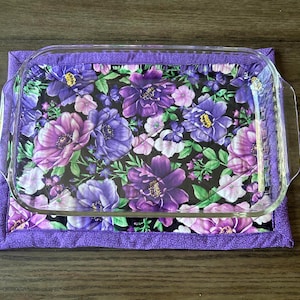 May include: A purple quilted hot pad with a floral print featuring purple, pink, and white flowers on a black background. A clear glass baking dish sits on top of the hot pad.