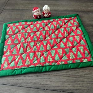 Pode incluir: Um jogo americano retangular de Natal com fundo vermelho e árvores de Natal verdes. O jogo americano tem uma borda verde e é acolchoado. Duas pequenas figuras de cerâmica do Papai Noel e da Mamãe Noel estão em cima.