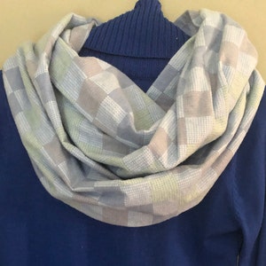 Puede incluir: Una bufanda suave y a cuadros en tonos de azul claro, gris y verde. La bufanda está doblada y drapeada alrededor de un suéter de cuello alto azul.