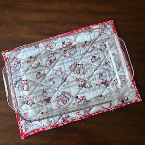 Puede incluir: Una almohadilla caliente acolchada blanca con un borde rojo y un patrón de pingüinos y muñecos de nieve. Un plato para hornear de vidrio transparente está encima de la almohadilla caliente.