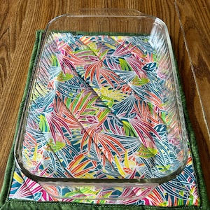 May include: A clear glass baking dish with a rippled edge sits on a colourful quilted hot pad with a tropical leaf pattern. The hot pad is green on the outside and has a white background with red, yellow, and green leaves.