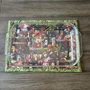Pode incluir: Um protetor de mesa acolchoado verde com um prato de vidro para assar em cima. O protetor de mesa tem uma estampa de frutas, garrafas de vinho e flores.