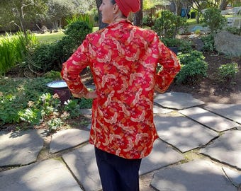 May include: A woman wearing a red and gold patterned long-sleeved jacket with a red turban. She is standing on a stone path in a garden.
