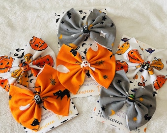 May include: Five fabric hair bows featuring Halloween-themed patterns including pumpkins, ghosts, bats, and stars in orange, gray, and white. Each bow is centered with a small character charm, such as a ghost, skull, or witch hat, and mounted on a card.