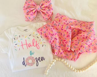 May include: A white short-sleeve bodysuit with the text Half a dozen and a donut graphic, paired with a pink skirt and a matching pink hair bow, both featuring a colorful sprinkle pattern. The bodysuit has the text 0-6 P. Custom written on it.