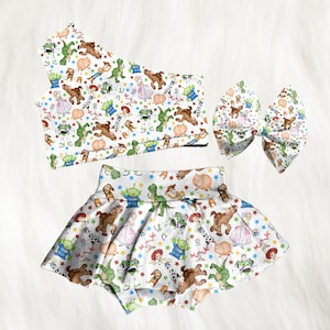 Può includere: Un completo per neonati composto da tre pezzi con sfondo bianco e fantasia colorata di personaggi dei cartoni animati. Il set comprende un top monospalla, una gonna e un grande fiocco. I personaggi includono un dinosauro, un maiale e un alieno.
