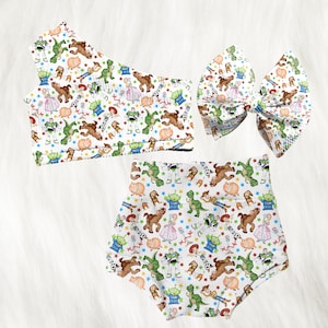 May include: A three-piece set featuring a white background with a colorful pattern of cartoon characters. The set includes a top, high-waisted bottoms, and a large bow. The characters include a dinosaur, a pig, and a cowboy.