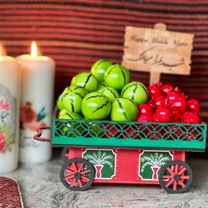 Può includere: Un carrello decorativo in legno pieno di oggetti verdi a forma di anguria e melograni rossi. Il carrello è rosso e verde con ruote nere. Due candele accese e un cartello di legno con "Happy Yalda Night" sullo sfondo.
