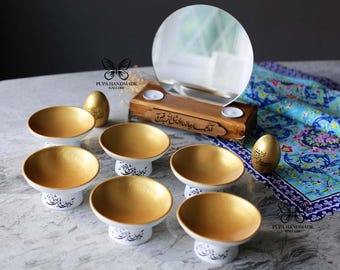 May include: A set of seven gold and white decorative bowls with Arabic calligraphy on the base. The bowls are arranged on a white surface with a wooden stand with a mirror and candles in the background. The stand has Arabic calligraphy on it.