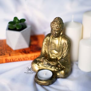 Op de afbeelding: Een gouden Boeddhabeeld staat op een wit oppervlak met een kleine witte kaars ervoor. Achter het Boeddhabeeld staan drie witte kaarsen in de vorm van een pilaar. Op de achtergrond is een boek met de titel "BHAGAVAD-GITA" te zien.