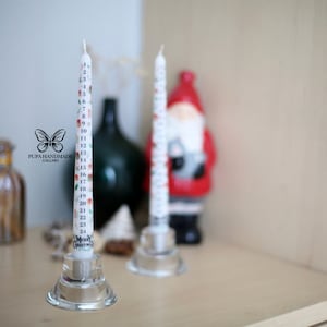 Può includere: Due candele d'avvento bianche con numeri stampati su di esse. Le candele sono su portacandele di vetro. La candela a sinistra ha il testo "Merry Christmas" stampato su di essa.