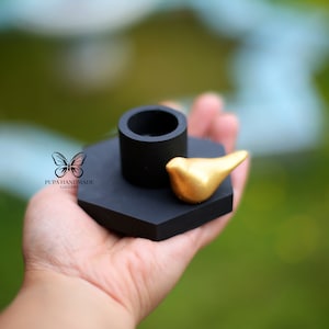 Puede incluir: Portavelas hexagonal negro con una pequeña figura de pájaro dorado en la parte superior.