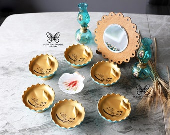 May include: A set of six teal and gold decorative bowls with Arabic calligraphy. The bowls are arranged in a circle on a gray surface. There are two teal glass oil lamps, a seashell, and a gold mirror with a teal frame in the background.