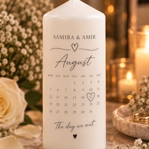 Op de afbeelding: Een witte cilindrische stompkaars met zwarte opdruk met de tekst SAMIRA & AMIR, August en The day we met. De kaars toont een kalender voor de maand augustus waarbij het getal 17 is omcirkeld door een hartvorm.