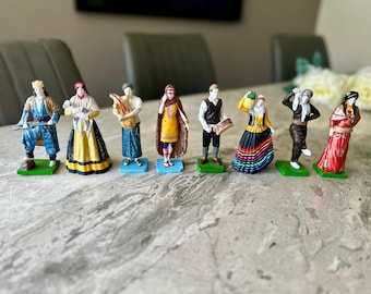 May include: A collection of eight miniature figurines depicting people in traditional attire. The figurines are painted with vibrant colors and stand on small green or blue bases. The figures represent various cultural styles.