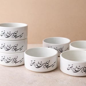 May include: A collection of white ceramic bowls with a cylindrical shape. Each bowl features black calligraphy around the exterior. Some bowls are stacked, while others are arranged separately.