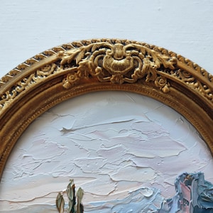 May include: An oval-shaped, ornate gold-colored frame with intricate relief carvings of leaves and scrolls along the top edge. The frame surrounds a textured oil painting featuring thick, visible brushstrokes in shades of pale pink, white, and blue.