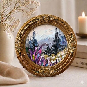May include: A circular, decorative gold frame holds a small landscape painting. The artwork shows a mountain scene with purple, pink, and green brushstrokes in the foreground, and blue and white mountains in the background. A lit candle and vase of flowers are in the background.